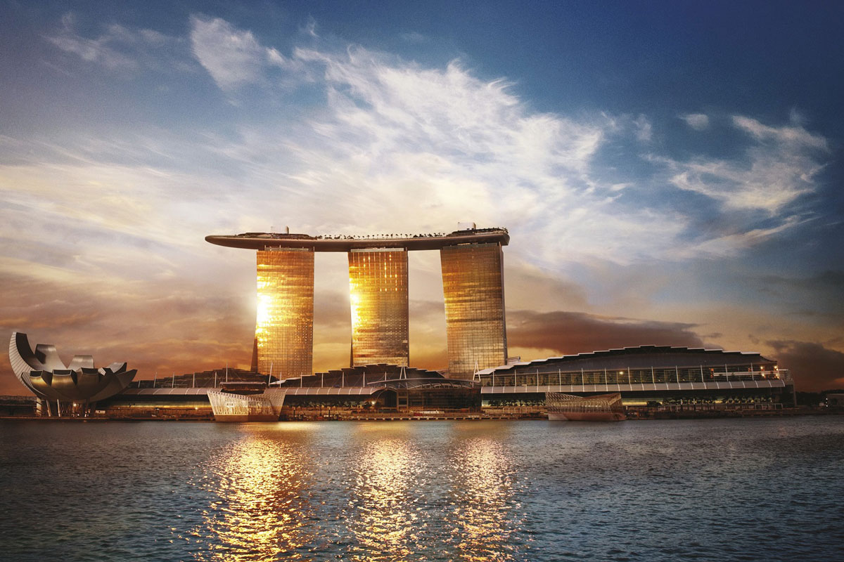 View of an the iconic Marina Bay Sand building which has three high-rise towers connected by a huge platform. The building is seen from across the water as the sun sets (or rises).